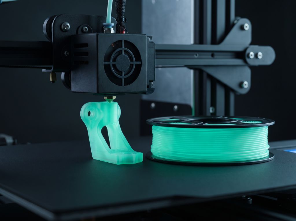 Düse eines 3D-Druckers über einem nachleuchtend gedruckten Bauteil, daneben eine Filamentrolle