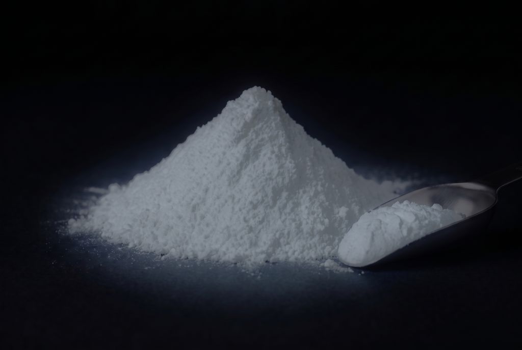 Weißes Nachleuchtpigment als Häufchen mit Messlöffel, im Dunkeln schwach weiß leuchtend